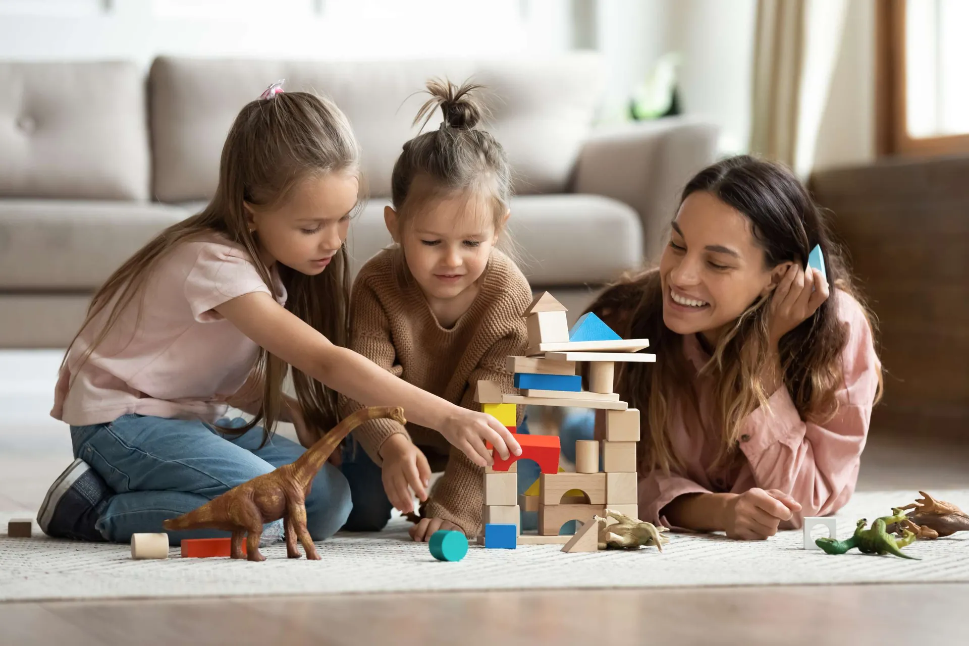 Caregiver building a block tower with two young girls on a living room rug